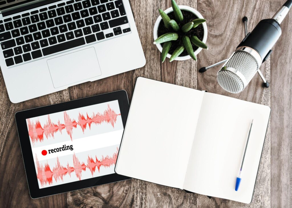 image of a laptop, ipad with sound waves and the word "recording", a microphone, a blank notebook and pen, and a plant