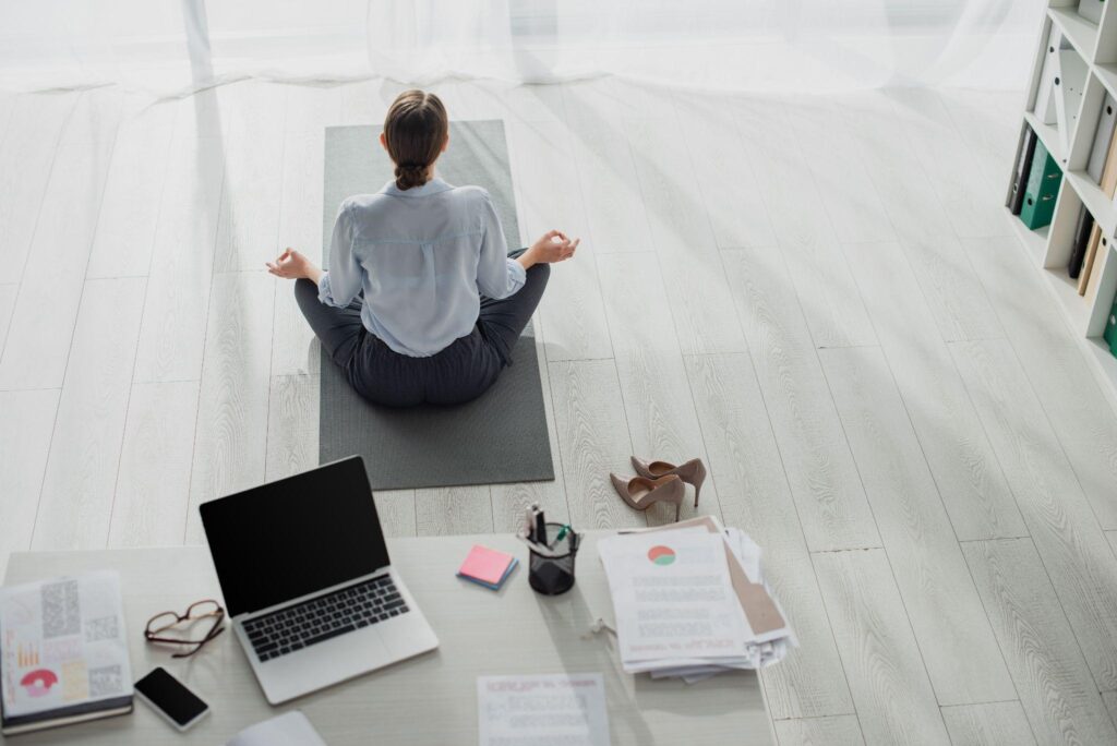 woman meditating in front of a desk with laptop, glasses, papers, phone, post-it notes, pencil cup