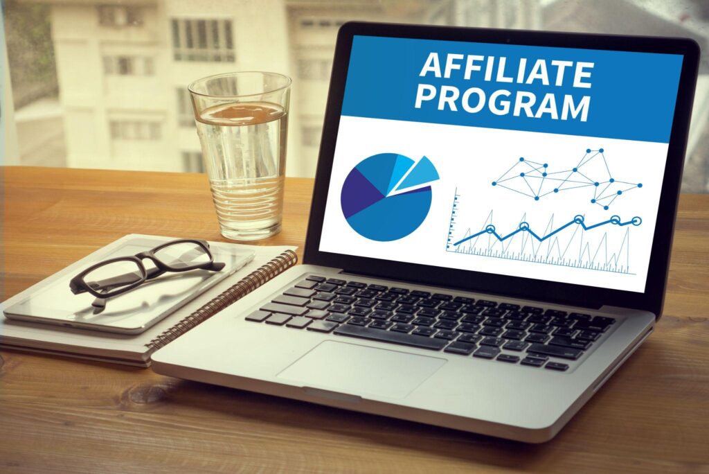 laptop with pie chart, graph, words "Affiliate Program" on table next to glass of water and notebook with eyeglasses on top