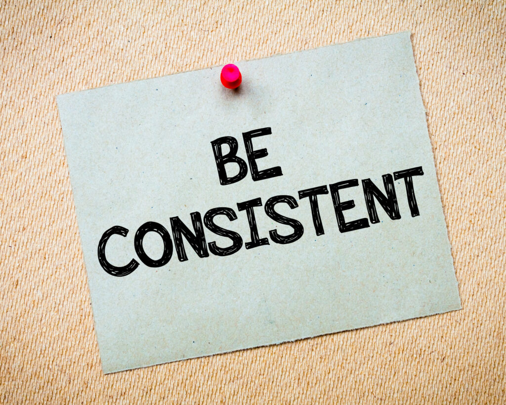 paper with the words "Be Consistent" written in black ink. Paper is pinned to brown background with a pink thumbtack