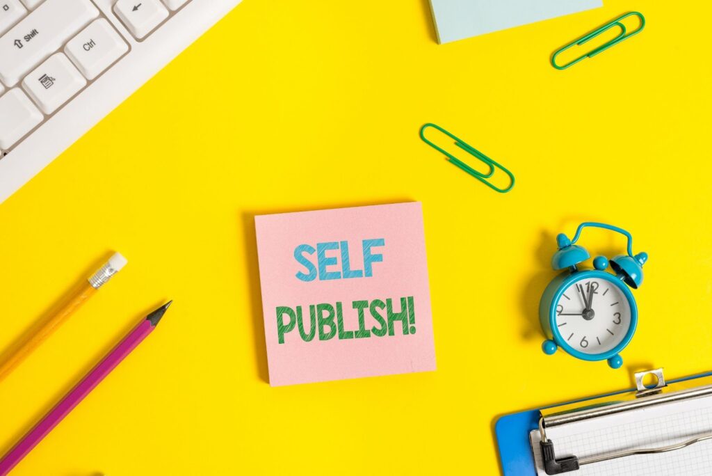 pink stick note with the words "Self Publish!" on it, surrounded by an alarm clock, paper clips, pencils, part of a keyboard, part of a clipboard