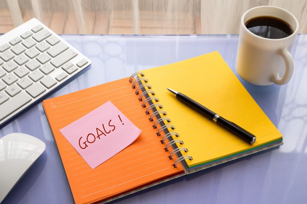 computer keyboard and mouse, cup of coffee, spiral notebook, pen, pink post-it note with "GOALS!" written on it