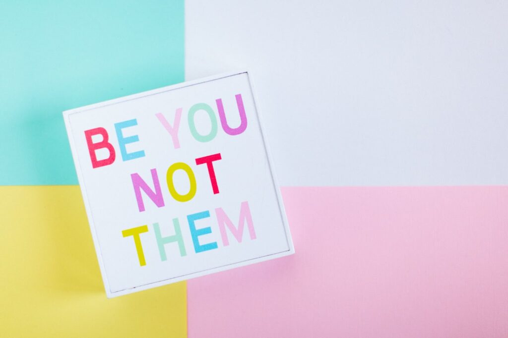 multi-colored background with a white sign with multi-colored lettering saying "Be You Not Them"