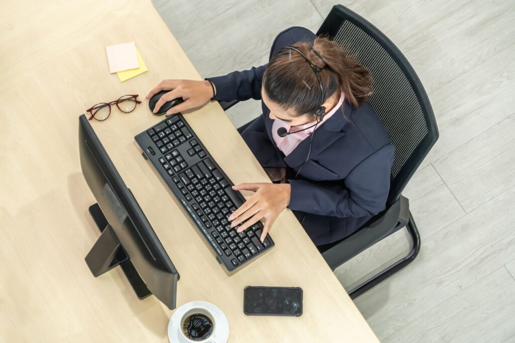 overhead view of a woman wearing a phone headset working on a computer. Cup of coffee, glasses, post-it notes, cell phone next to her