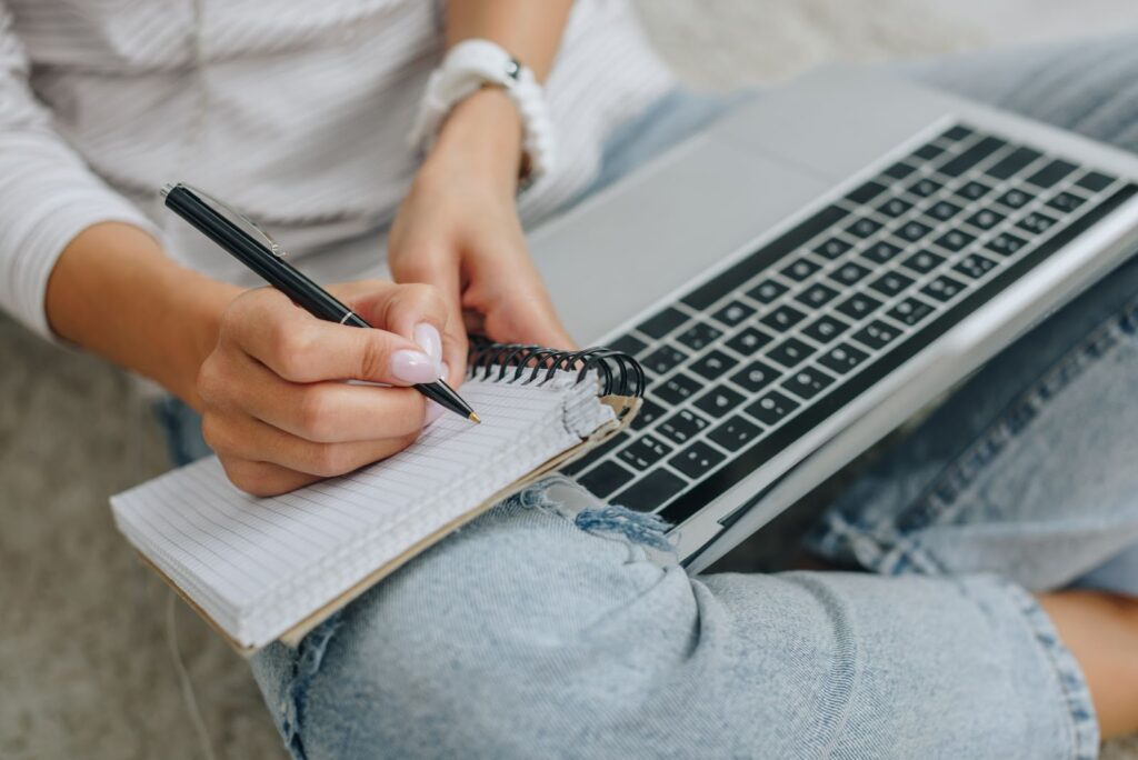 woman (shown from waist down) sitting on the floor with a laptop in her lap and writing on a notepad