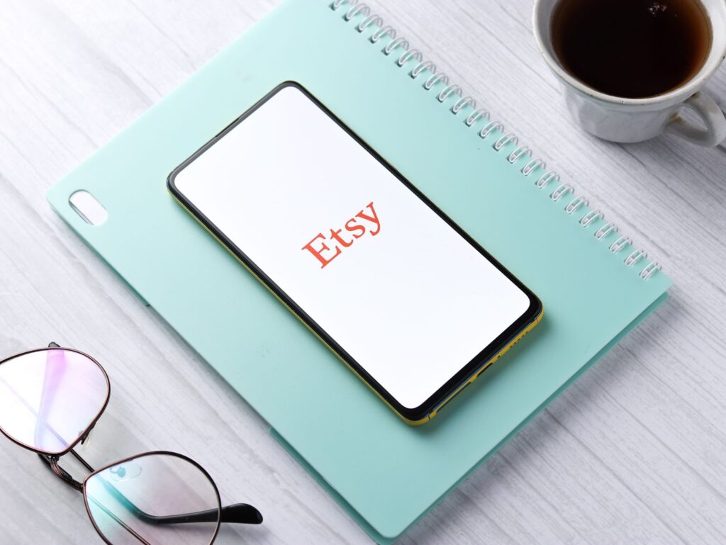 light teal notebook with a cell phone on top. Etsy logo on the phone screen. Eyeglasses and cup of coffee on either side of the notefook.