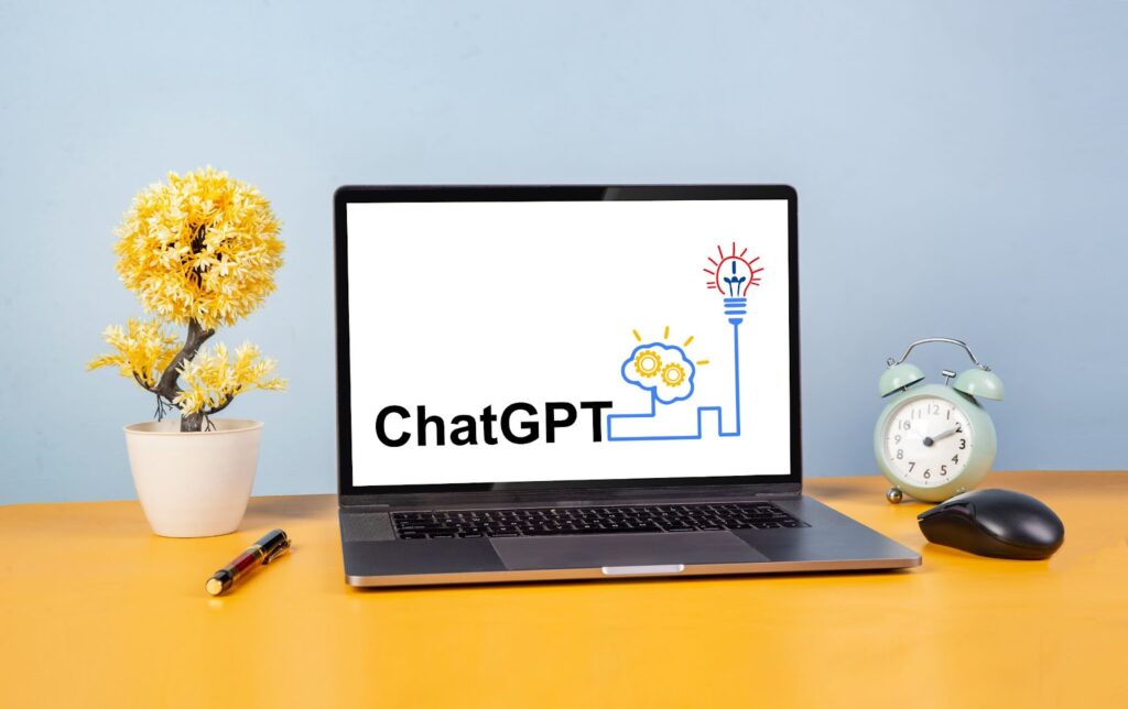laptop computer with "ChatGPT" text and stylized drawing of brain connected to lightbulb on screen. Pen and plant with yellow leaves on table to left of laptop; alarm clock and computer mouse to right of laptop