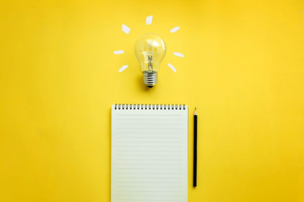 blank top-spiral notebook with pencil to the right; lightbulb above notebook; bright yellow background