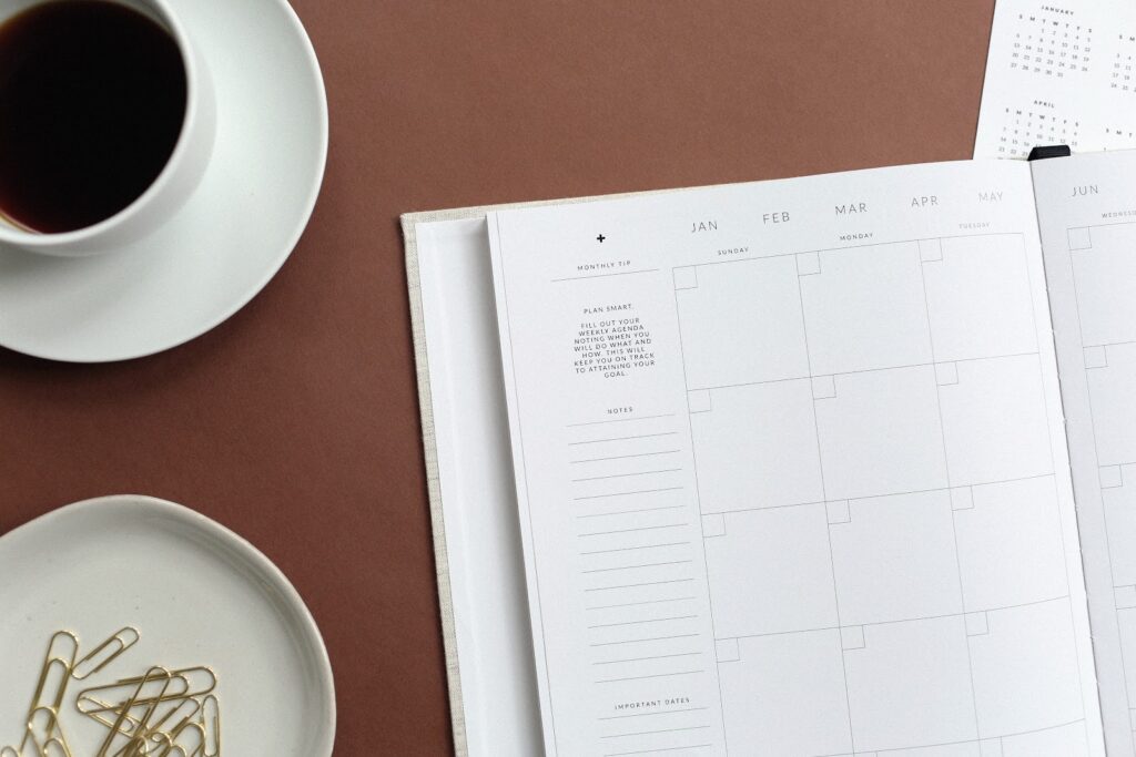 open, blank planner on a table. cup of coffee on a saucer in upper left, tray with gold paper clips in lower left