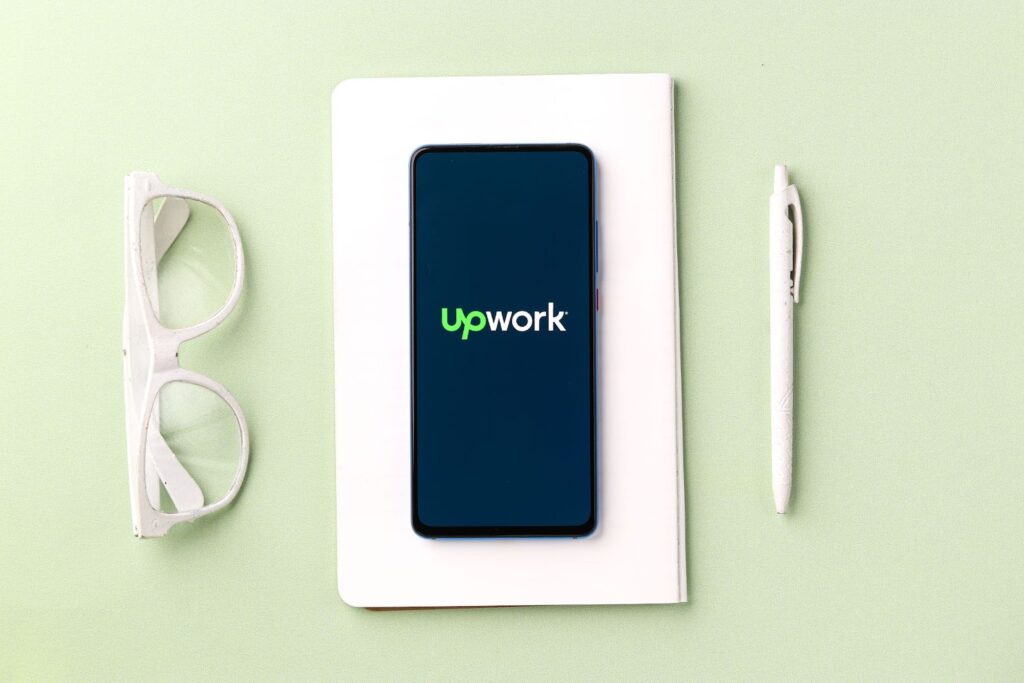 cell phone with Upwork logo on the screen. Phone sitting on a blank journal page. Eyeglasses on the left side, pen on right side