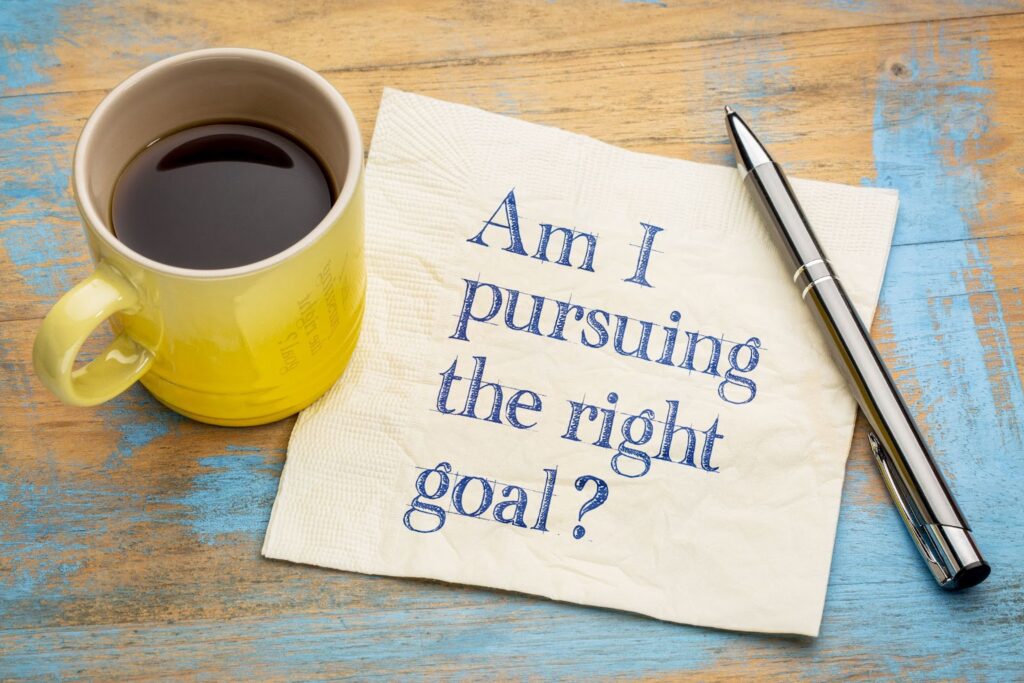 Words "Am I pursuing the right goal?" written on a napkin. Pen and cup of coffee sitting next to napkin.