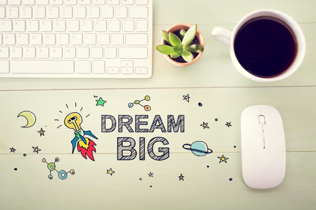 text "Dream Big" surrounded by drawings of planets, stars, rocketship; on table -- computer keyboard, cactus plant, cup of coffee, computer mouse