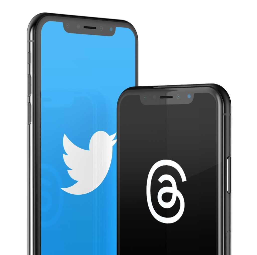 two cell phones; phone on left displaying the twitter logo on a blue screen; phone on the right displaying the Instagram Threads logo on a black screen