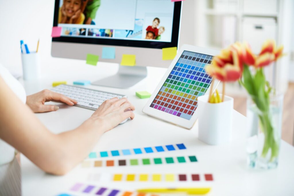 woman working at computer; ipad with color swatches next to her; paper color swatches on desk next to her; pencil cup and vase of flowers on desk