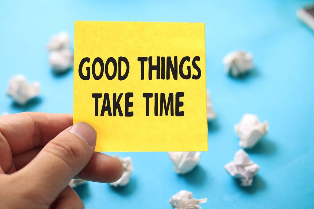 hand holding yellow post-it note with text "Good things take time"; blue background with crumpled up pieces of paper
