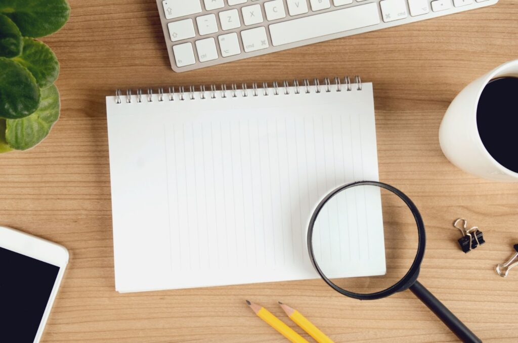 items on a desk: blank spiral notepad, magnifying glass, pencils, binder clips, cup of coffee, computer keyboard, ipad, green leafy plant